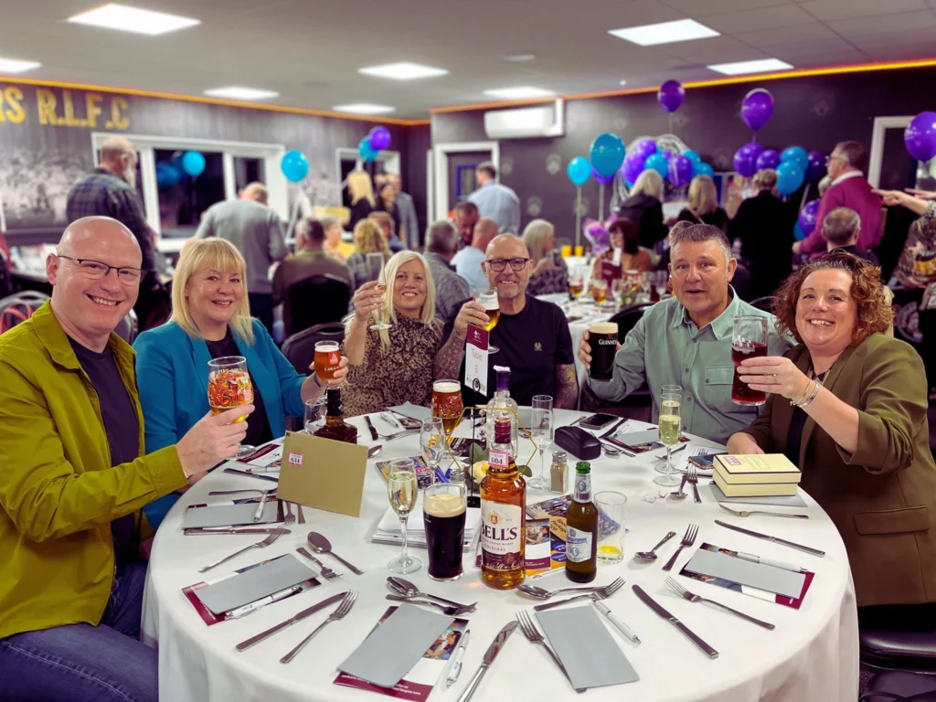 A group of six people sitting at a round table in a decorated function room, smiling and raising their glasses for a toast.