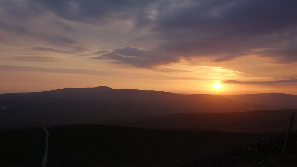 A wide landscape view of the Yorkshire 3 Peaks at sunset, showing the silhouette of rolling hills under a sky of orange and purple clouds.