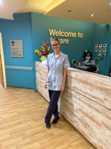 A smiling woman in a light blue healthcare tunic and dark trousers standing in front of a wooden reception desk with a teal wall background.