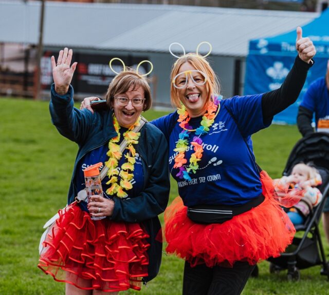 Two women smiling at an outdoor event, wearing red tutus, flower leis, and neon headbands. One waves while the other gives a thumbs up.