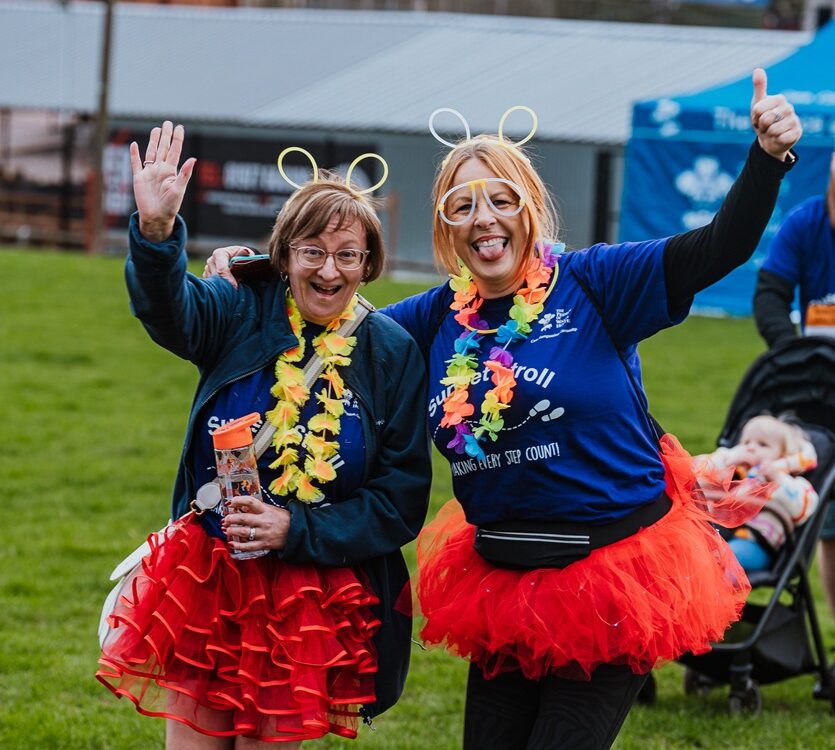 Two women smiling at an outdoor event, wearing red tutus, flower leis, and neon headbands. One waves while the other gives a thumbs up.