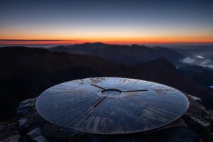 A view from the summit of Yr Wyddfa (Snowdon) at sunrise, featuring a metal mountain compass dial in the foreground and the silhouettes of mountain ranges against an orange and blue horizon.