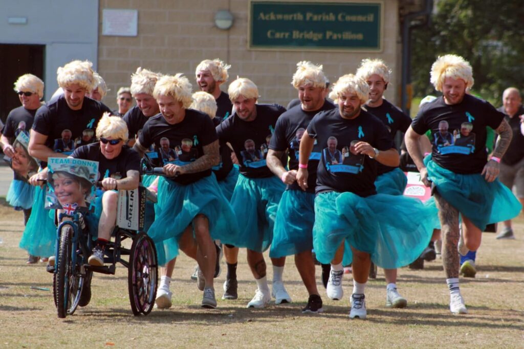 A group of men wearing black T shirt sand Blue Tutus with blond wigs run with 2 of them pushing a man in the same clothes on a bike.