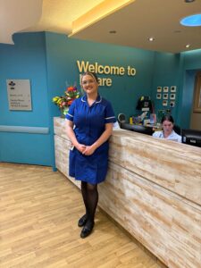 A smiling woman in a blue healthcare uniform standing in front of a wooden reception desk. Behind her, a teal wall features the text "Welcome to Care" and a vase of colorful flowers.A smiling woman in a blue healthcare uniform standing in front of a wooden reception desk. Behind her, a teal wall features the text "Welcome to Incare" and a vase of colorful flowers.