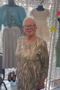 A woman with short white hair and glasses wearing a green and white patterned top, standing in front of a clothing display featuring a blue blouse and a white dress.