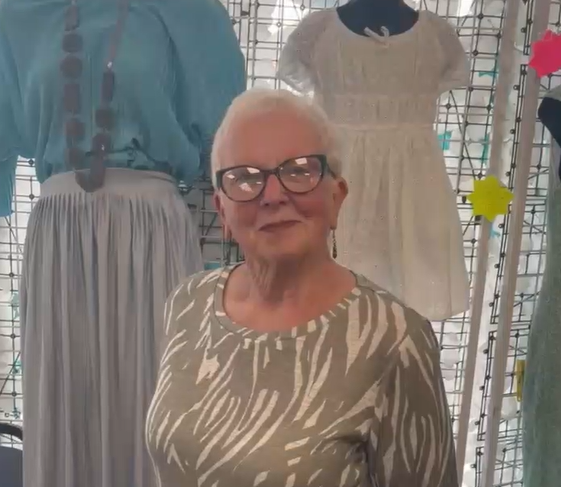 A woman with short white hair and glasses wearing a green and white patterned top, standing in front of a clothing display featuring a blue blouse and a white dress.
