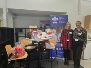 Two women standing next to a Prince of Wales Hospice banner and a table filled with various gift hampers and raffle prizes.