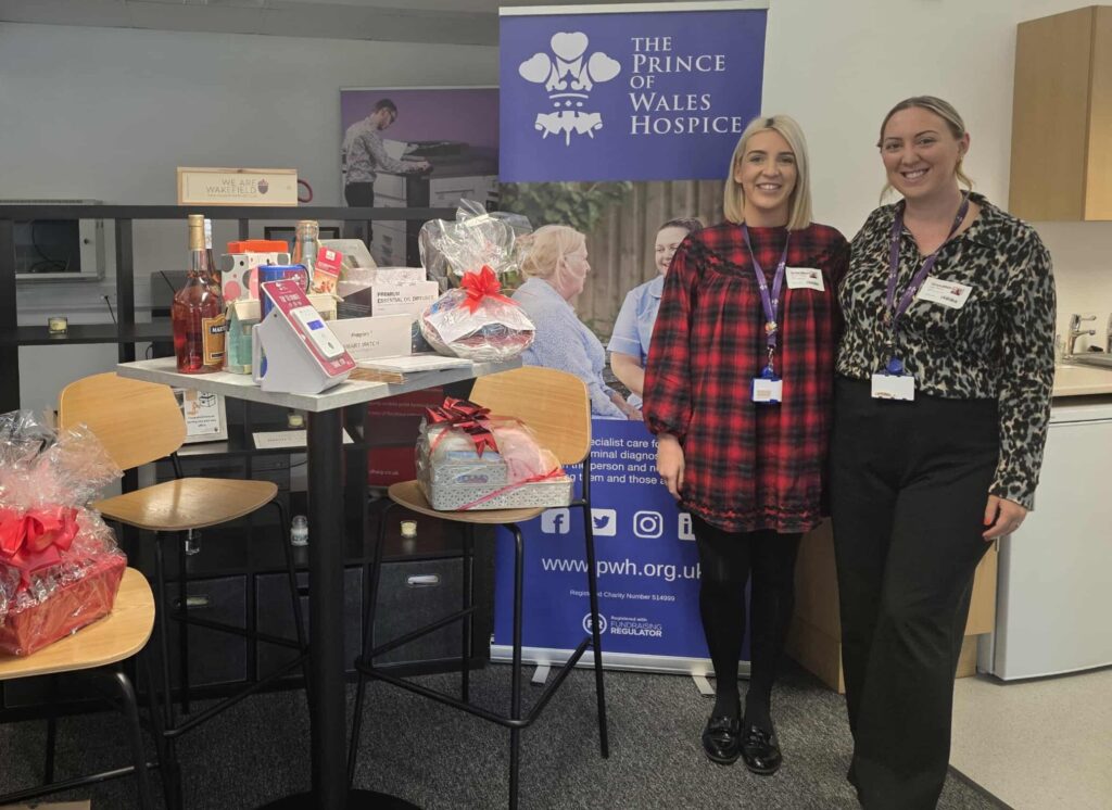 Two women standing next to a Prince of Wales Hospice banner and a table filled with various gift hampers and raffle prizes.