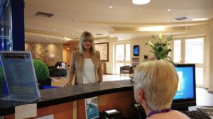 A lady with long blond hair is standing at the reception of the Hospice and talking to the receptionist.