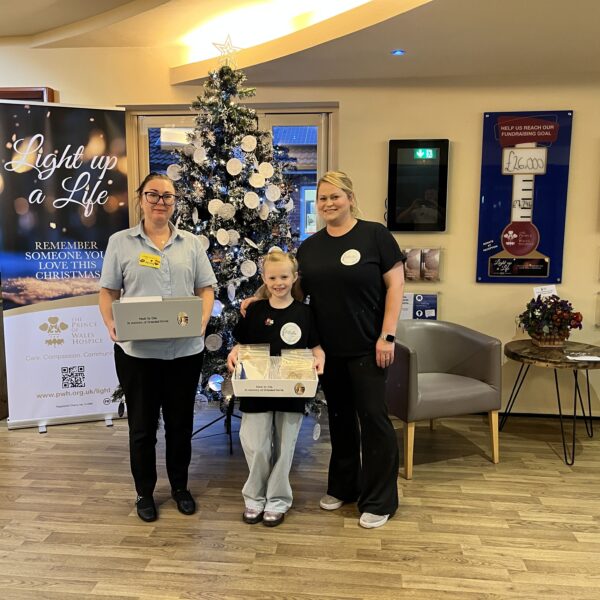 Two women and a young girl pose with handmade boxes beside a decorated Christmas tree and “Light up a Life” hospice fundraising displays.