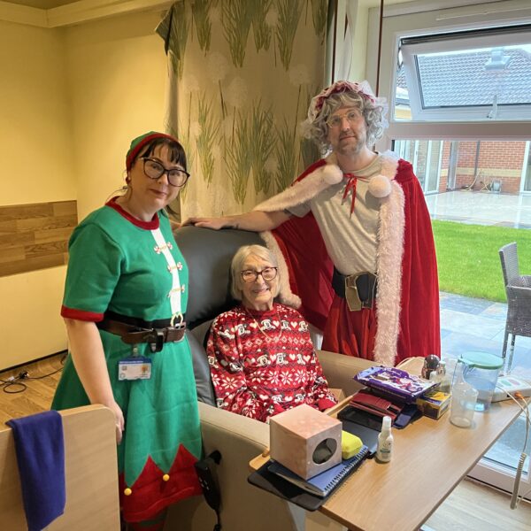 Three people, including someone dressed as Santa and someone dressed as an elf, gather around an elderly person in a hospital bed while holding and displaying chocolate packages.