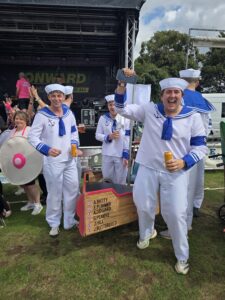 People in sailor costumes pose with drinks in front of an outdoor event stage, beside a wooden sign listing names.