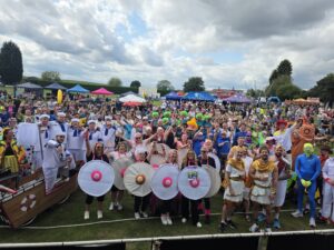 Large costumed crowd posing at an outdoor festival with vendor tents and inflatable attractions under a cloudy sky.