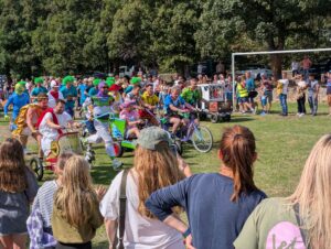 Crowded outdoor event with costumed participants riding colorful bicycles and pedal vehicles while spectators watch from a grassy field.