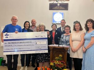 Ten people pose indoors with an oversized £1,551 cheque payable to The Prince of Wales Hospice.