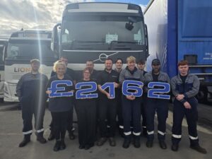 A group of 11 people poses outdoors in front of several large Volvo trucks. Most of the people wear dark work uniforms with reflective bands, while several hold oversized blue-and-white characters spelling “£2162.”