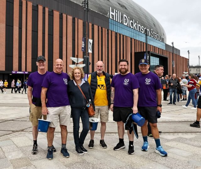 Six adults pose outside Hill Dickinson Stadium, with the stadium façade and crowds visible behind them.