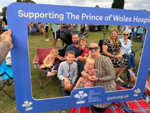 Family group posing behind a large blue Prince of Wales Hospice event frame on a grassy lawn.