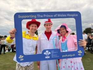 Three smiling people in colorful costumes pose behind a blue frame supporting The Prince of Wales Hospice.