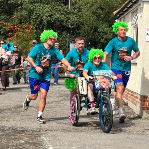 Runners in teal shirts and neon-green wigs accompany a person in a decorated bike along a street.