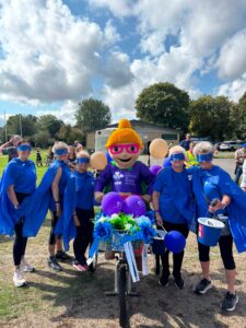 People in blue capes and masks pose with a costumed mascot and a balloon-decorated cart at an outdoor event.
