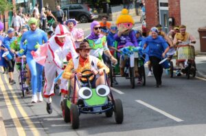 Costumed parade participants push decorated carts and vehicles along a city street, including a green cartoon-faced vehicle in the foreground.