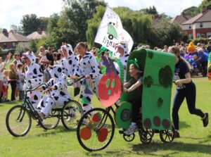 participants in cow costumes ride decorated bicycles alongside a large green caterpillar float while spectators watch.