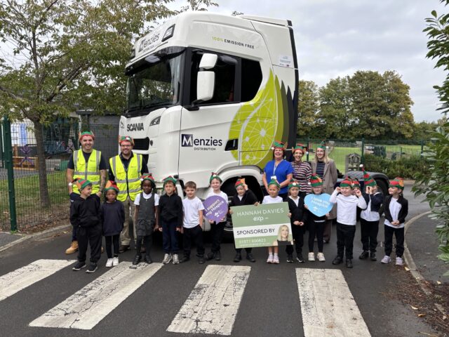 Children and adults in elf hats pose with hospice-support signs in front of a Menzies Distribution truck.