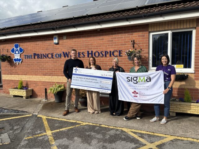 Five people outside The Prince of Wales Hospice hold a £10,000 presentation cheque and a Sigma fundraising banner.