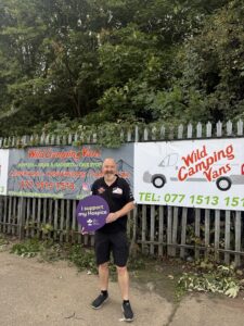 A man holding an “I support my Hospice” sign stands in front of Wild Camping Vans advertising banners and dense greenery.