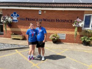 Two people wearing blue shirts and medals pose outside The Prince of Wales Hospice, beneath its crown logo.