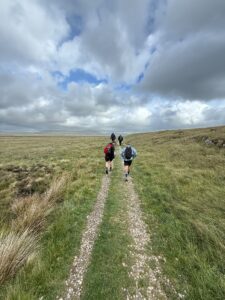 Four hikers walk along a rocky trail through grassy hills beneath a dramatic cloudy sky.