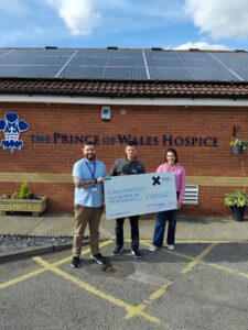 Three adults hold a large donation cheque outside the Prince of Wales Hospice, beneath rooftop solar panels.