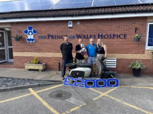 Four adults and a small dog pose beside a vintage scooter outside The Prince of Wales Hospice, with a large “£2000” graphic in the foreground.